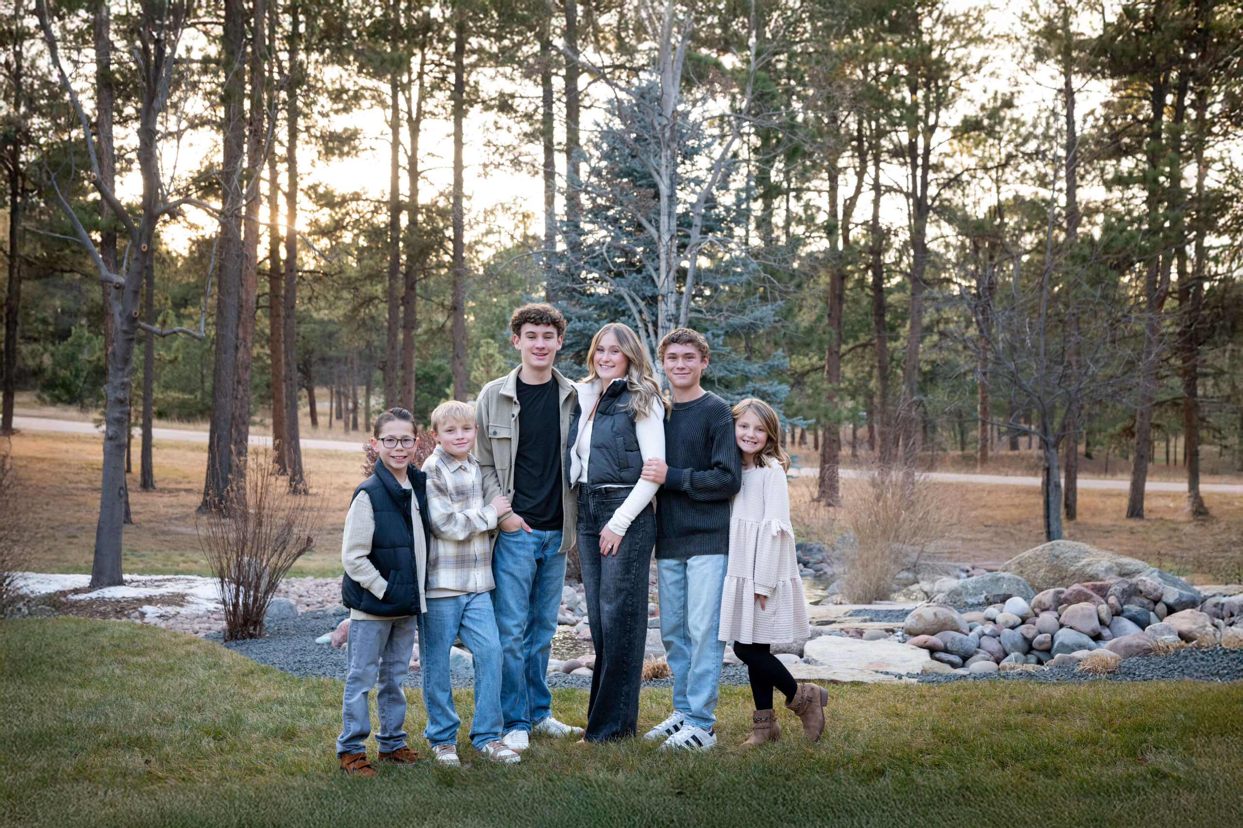Six happy siblings in jeans and tan stand in a pine forest at sunset after finding great indoor playgrounds in colorado springs