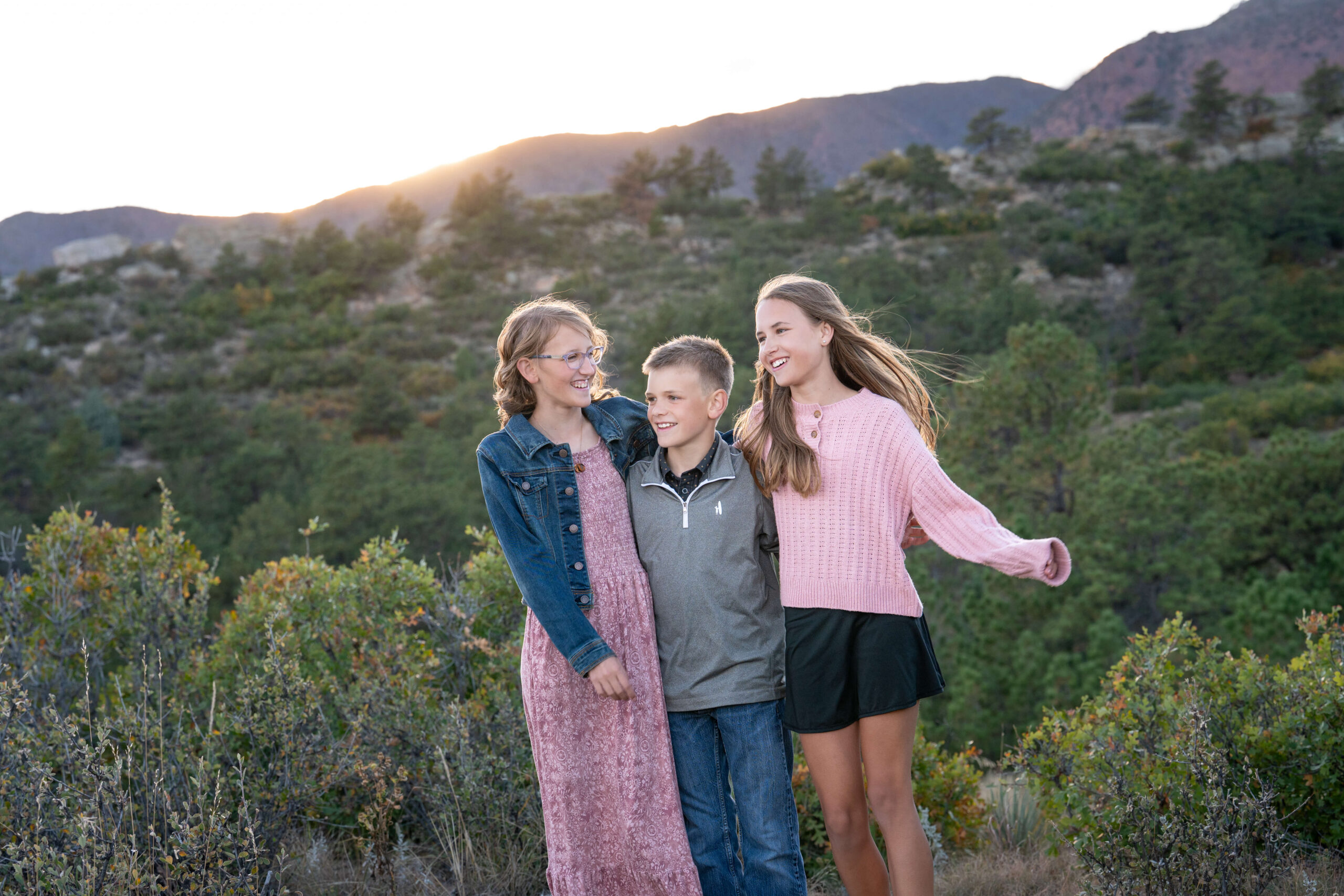 Three young siblings laugh while walking a park trail with arms around each other at sunset