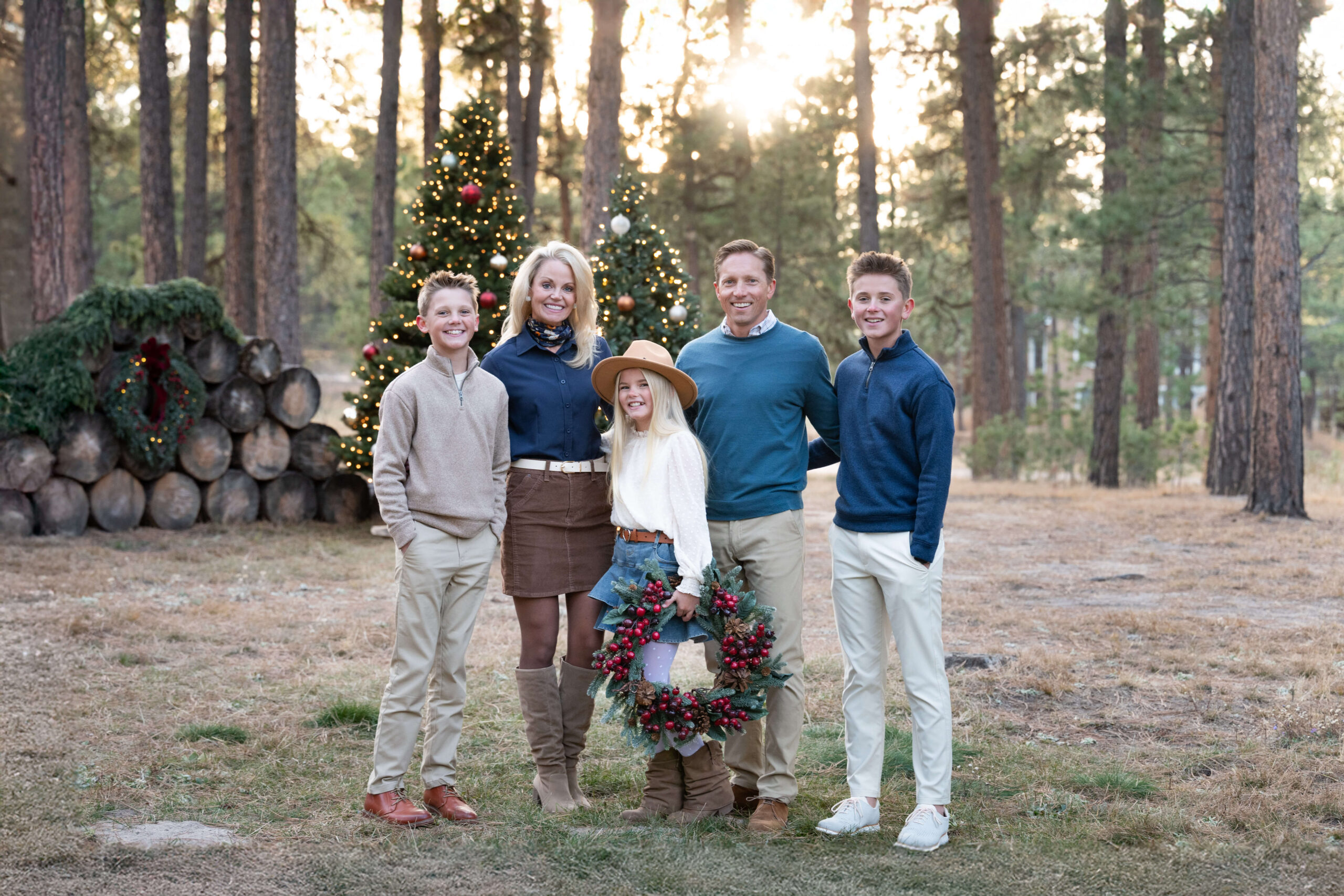 A mom and dad stand with christmas trees and their young daughter holding a wreath between their two teen sons at sunset after using montessori schools in colorado springs