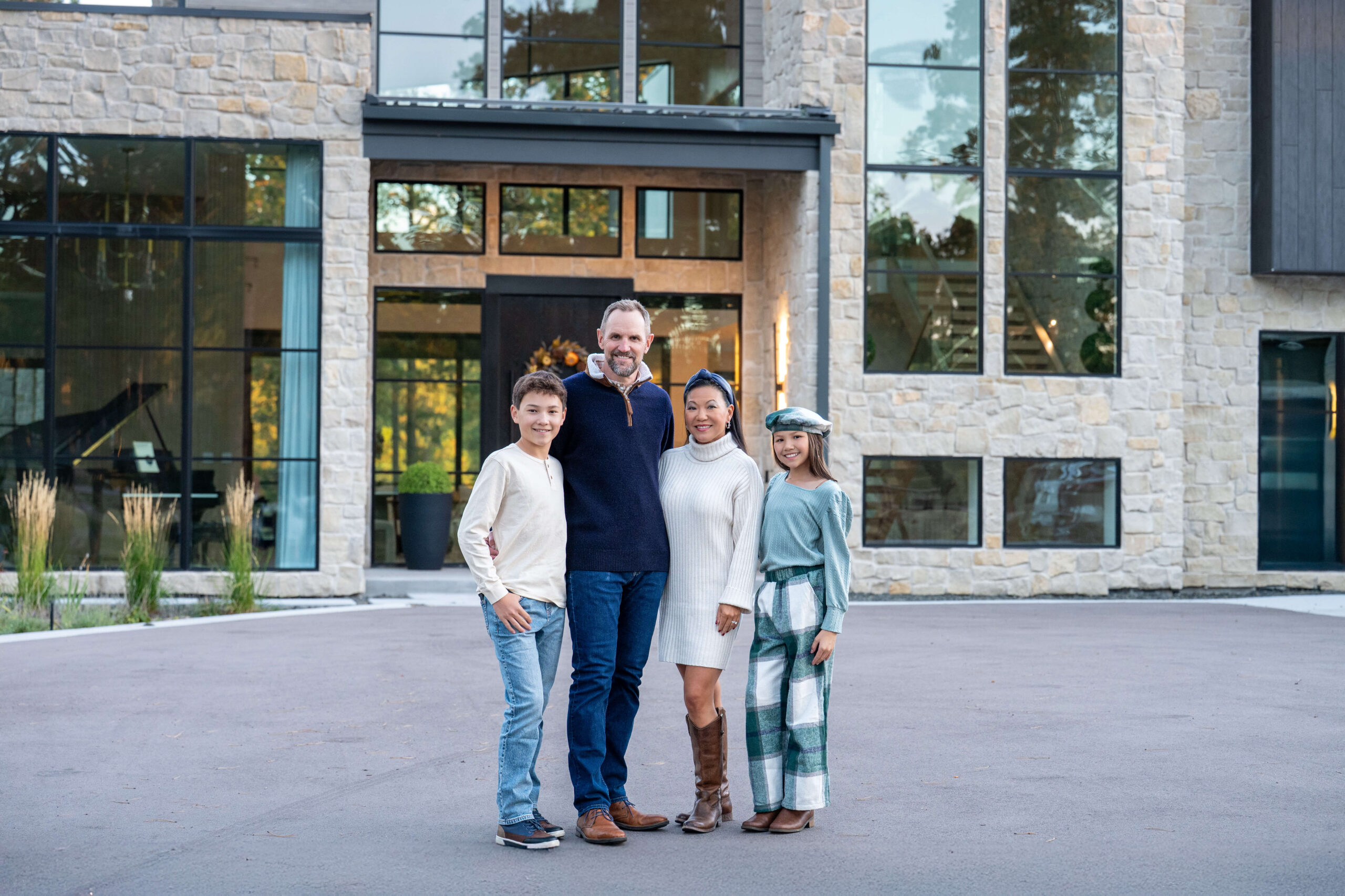 Happy mom and dad stand in a driveway of a modern luxury home with their two young children