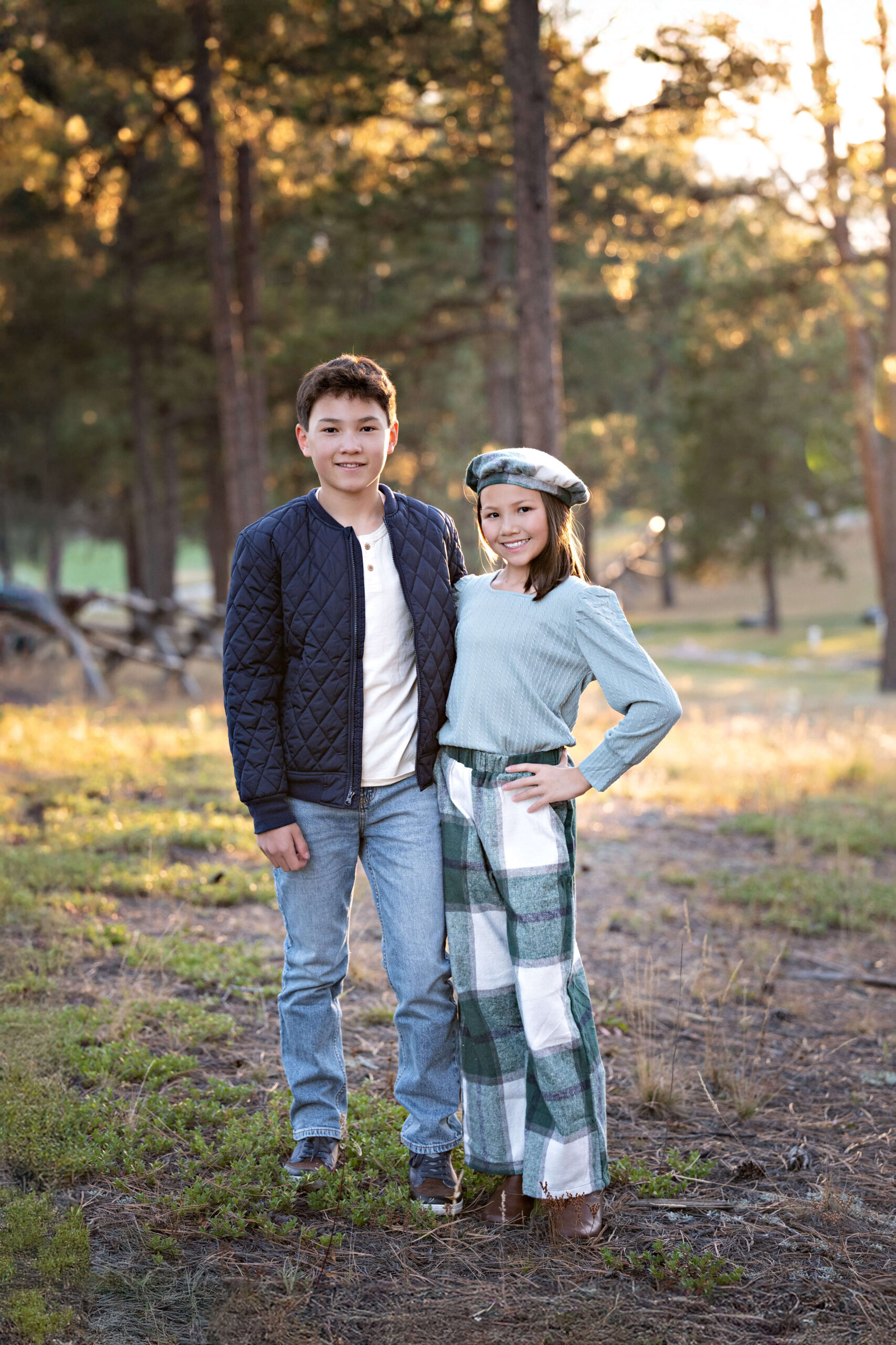 Young brother and sister in blue and green standing in a pine forest at sunset smiling with arms around each other after finding a great pediatric dentistry in colorado springs