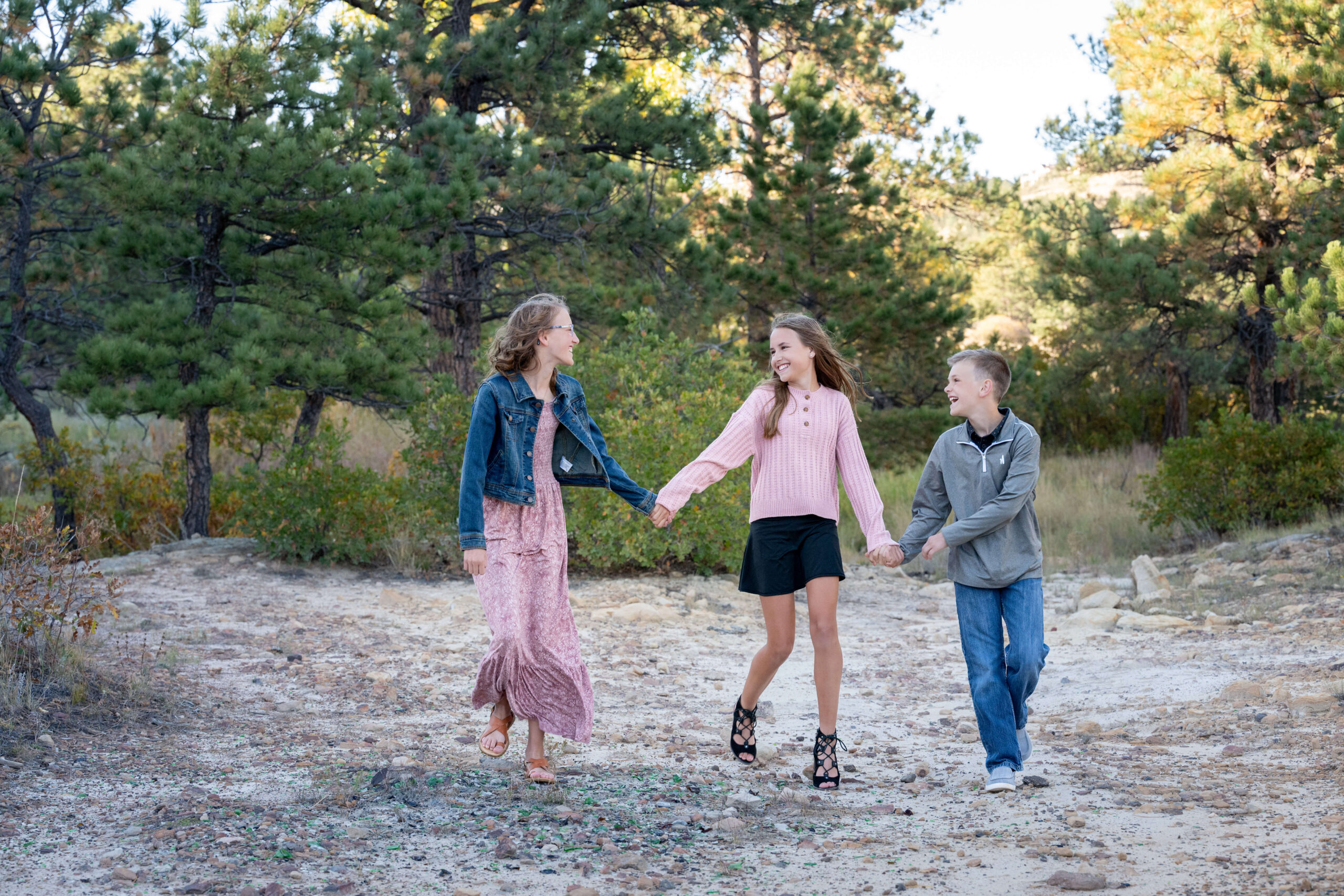 Three young siblings walk laughing and holding hands in a pine forest trail at sunset in pink and blue