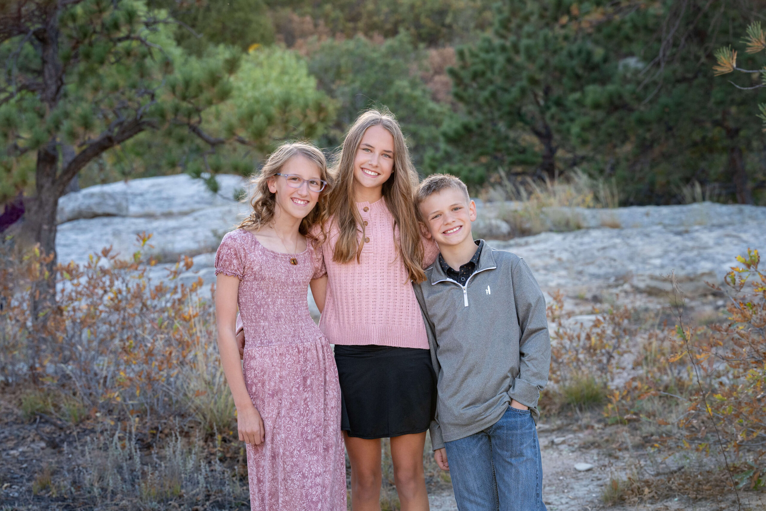 Three young siblings smiling with arms around each other in a mountain trail at sunset after finding pediatricians in colorado springs