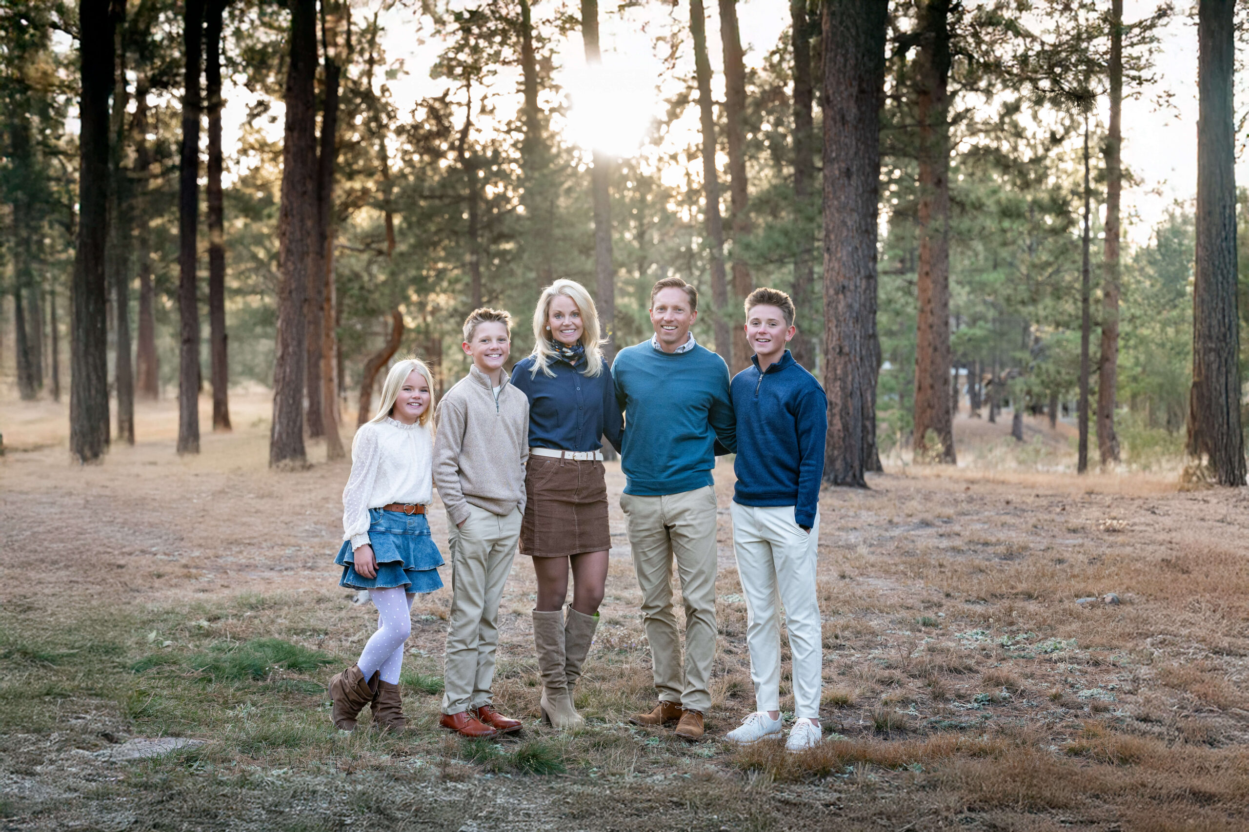 A mom and dad stand with their three young children in a pine forest at sunset smiling with arms around each other