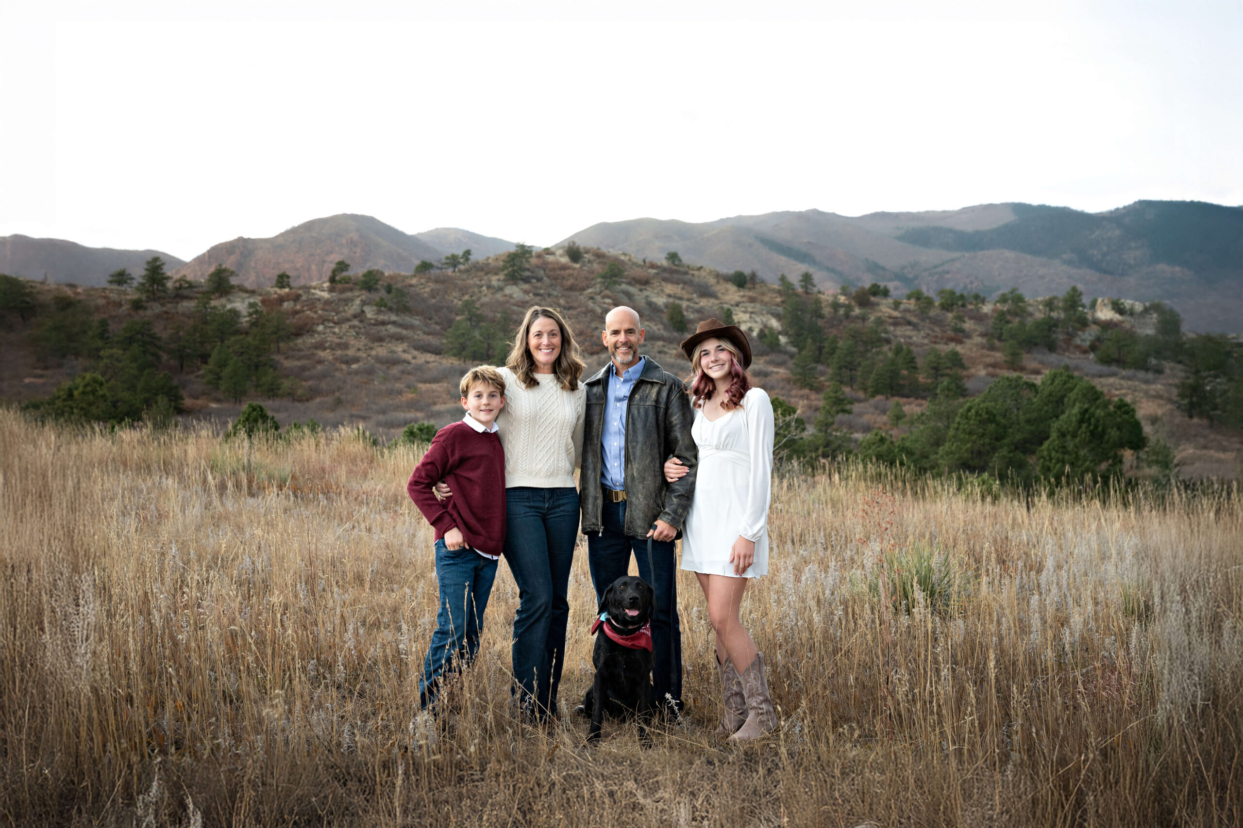 A mom and dad stand with their teen son and daughter and black dog in a mountain trail lined in tall golden grass at sunset after finding playgrounds in colorado springs