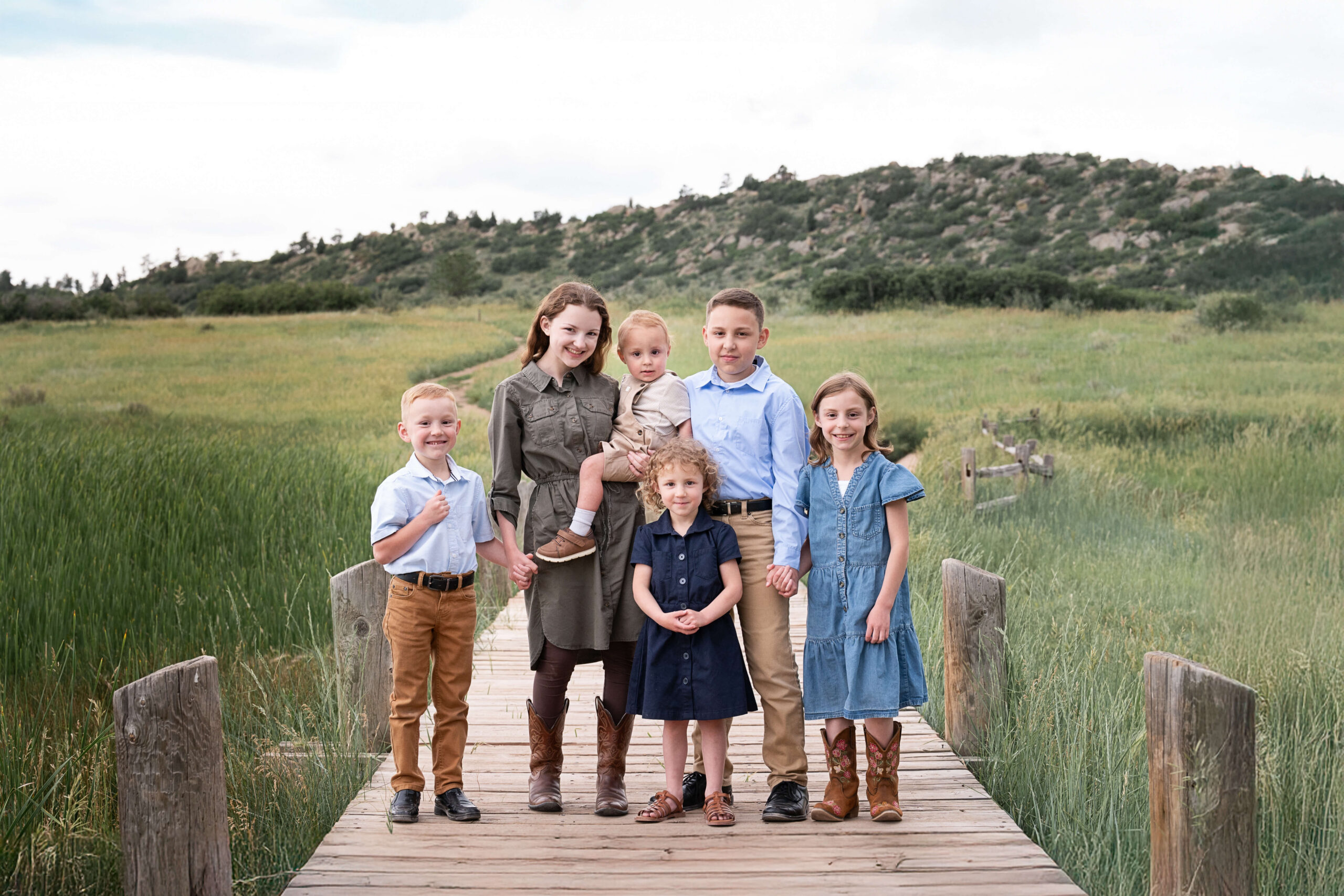 six young siblings stand smiling on a small boardwalk in a trail at sunset holding hands