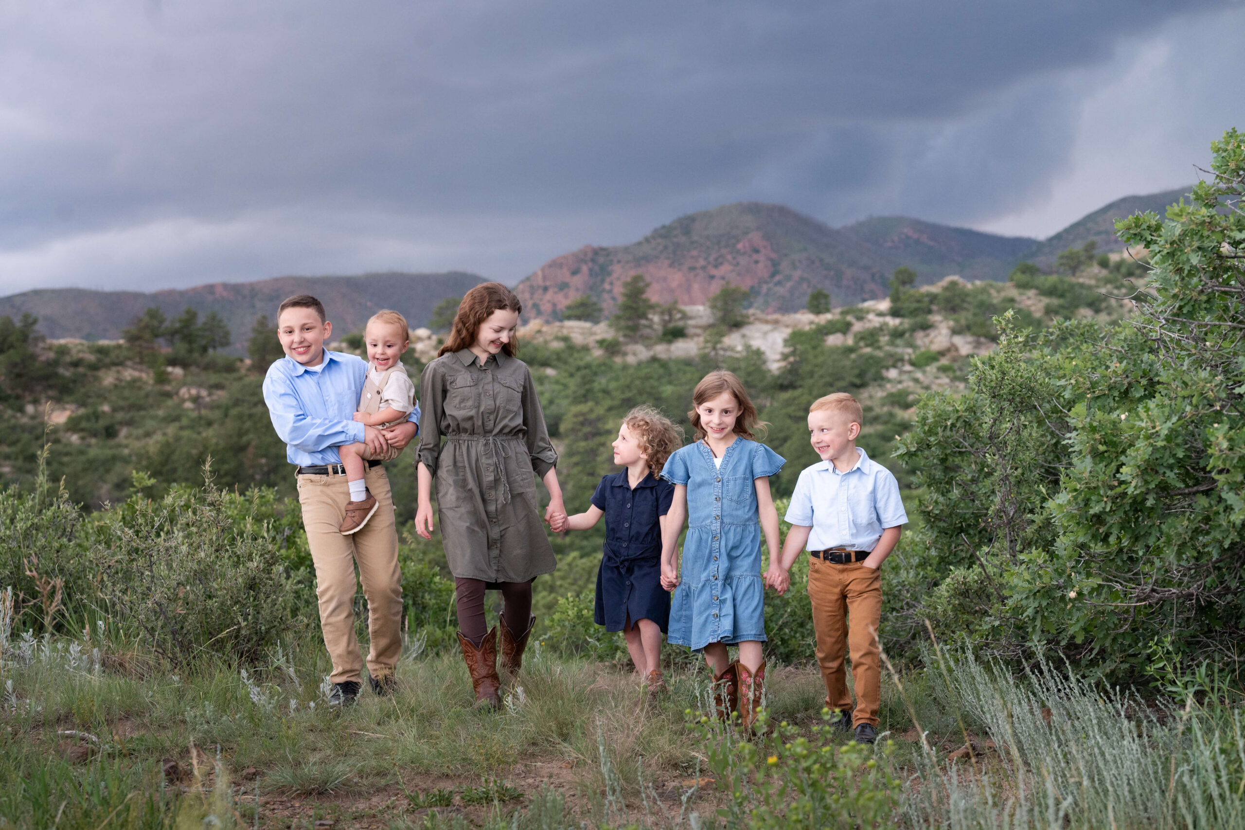 Six siblings under 12 walk holding hands and smiling in a park trail on an overcast day with orange mountains in the distance before visiting splash pads in colorado springs