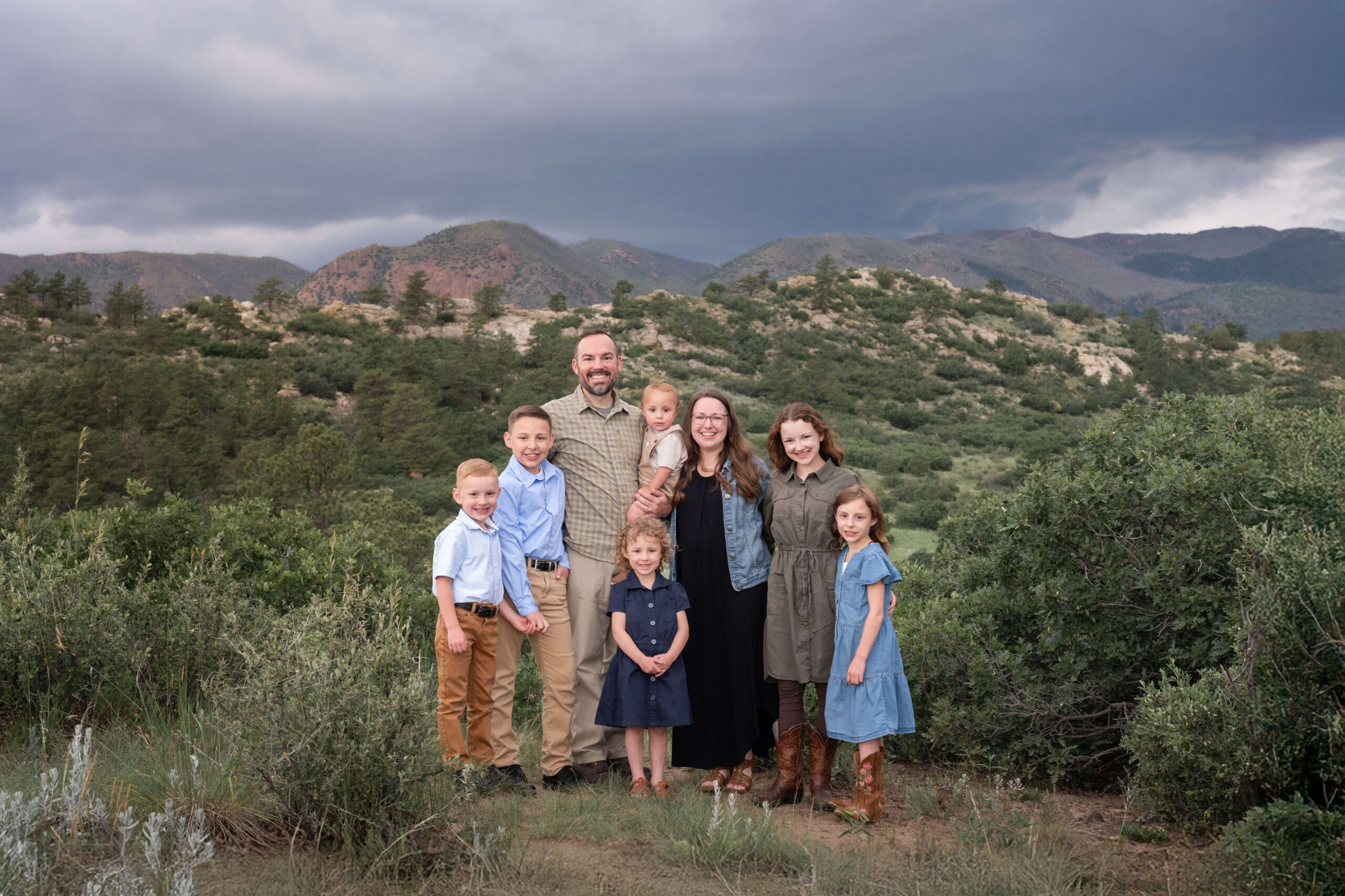 A smiling mom and dad standing in a mountain trail with their six happy children on an overcast day before visiting splash pads in colorado springs