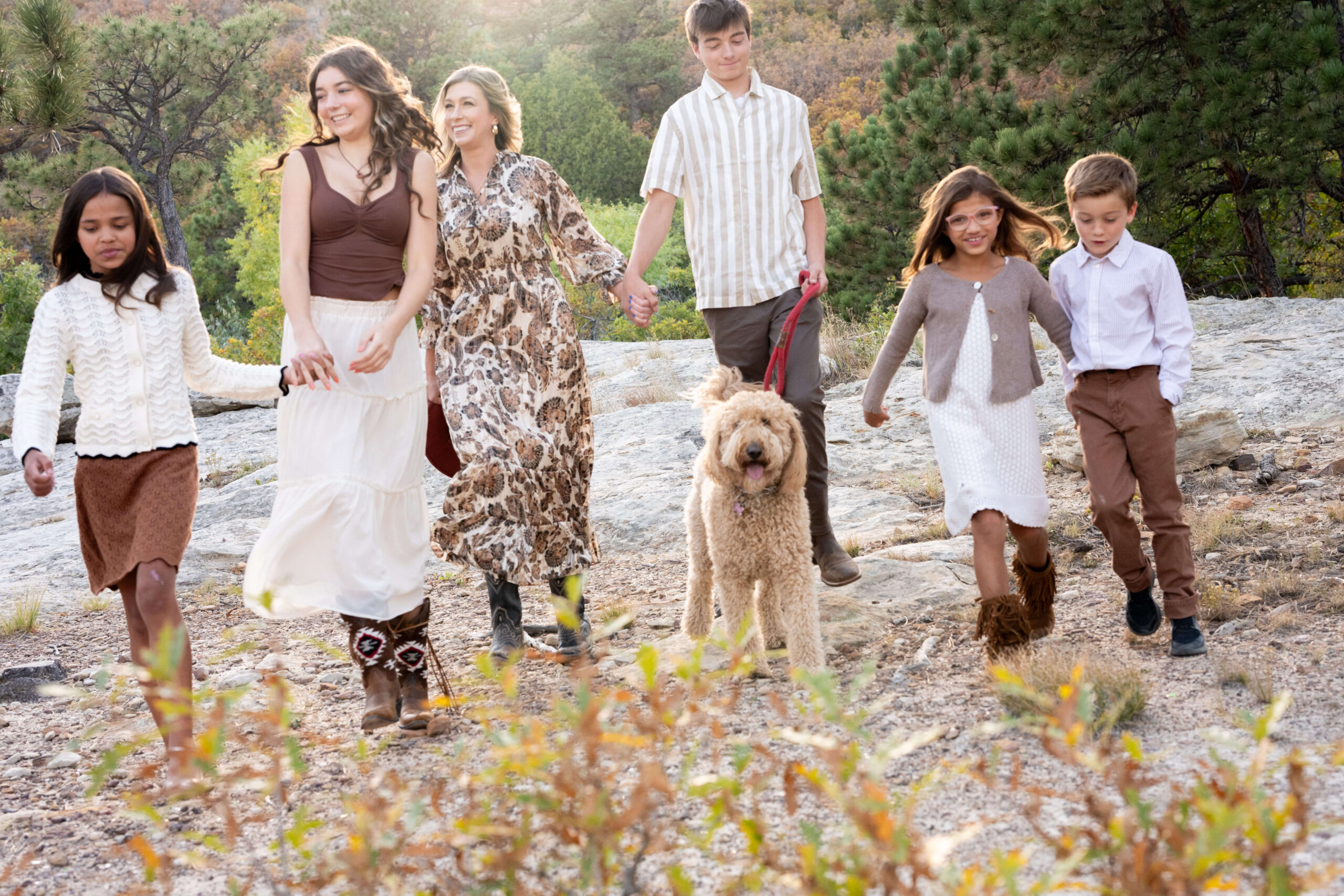 A happy mother walks with her 5 children and golden doodle on a mountain trail at sunset while holding hands