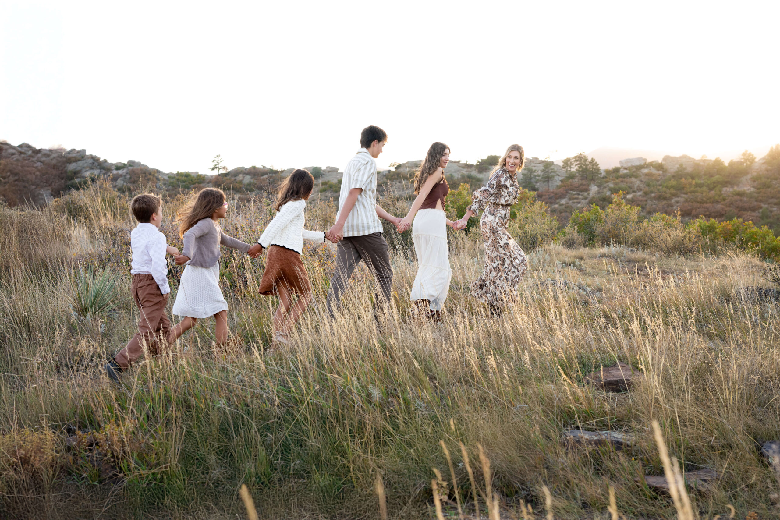 A mom looks back while running through a trail of tall golden grass at sunset with her five kids holding hands at sunset after finding summer camps in colorado springs
