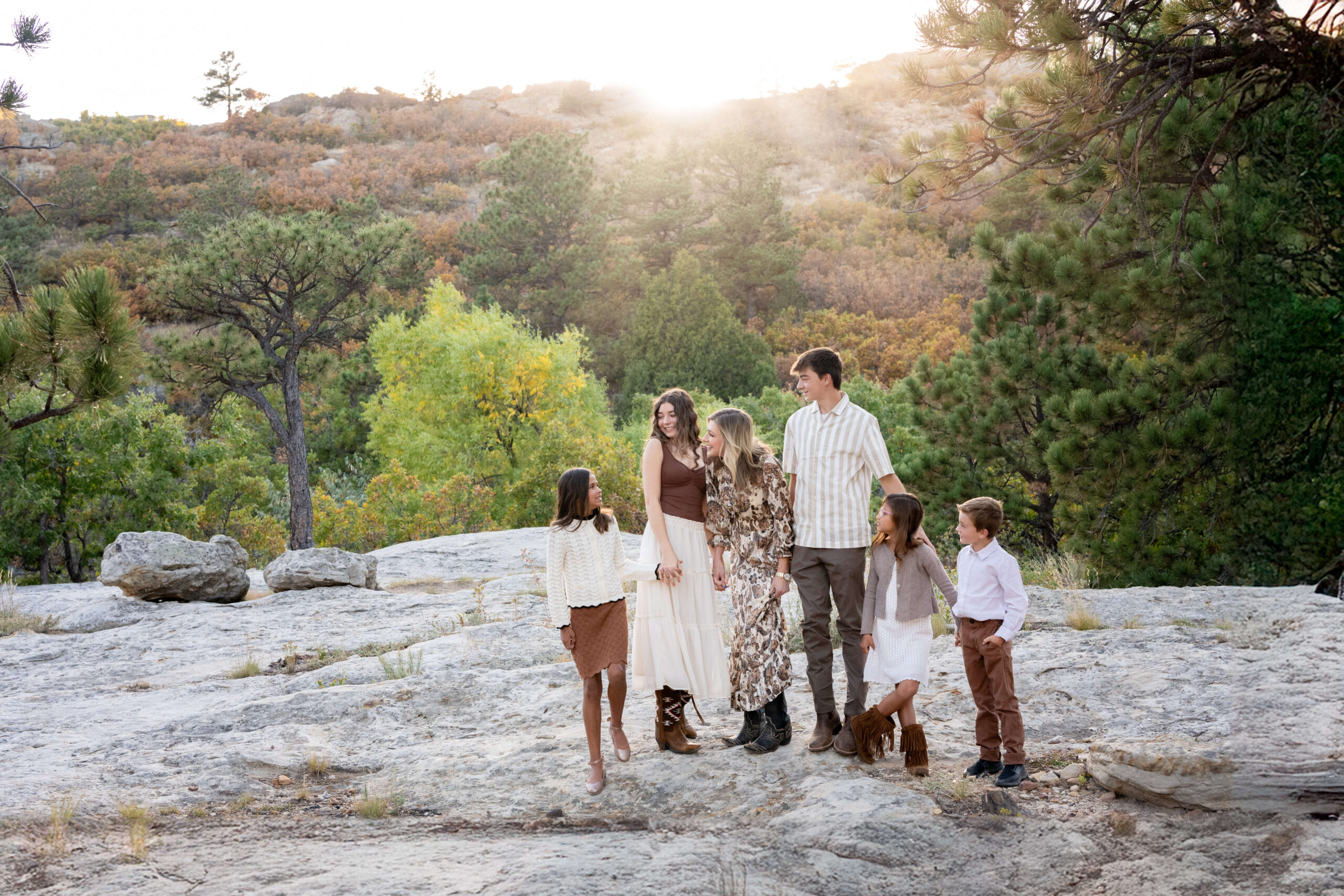 A mom stands on a mountain at sunset playing with her five young and teen children in white and tan after exploring summer camps in colorado springs