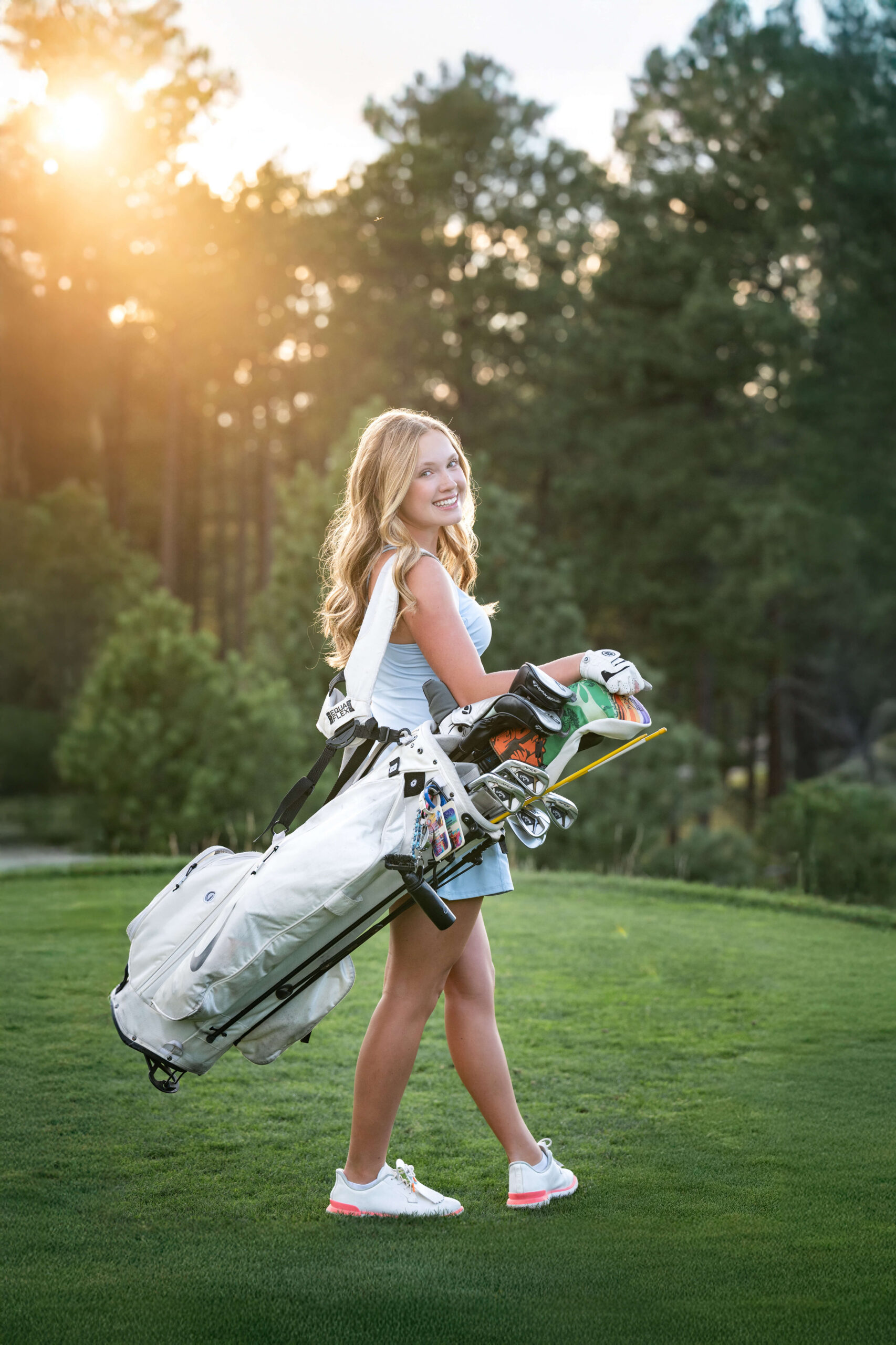 A high school senior walks on a putting green with her golf club carrier on her shoulder at sunset.