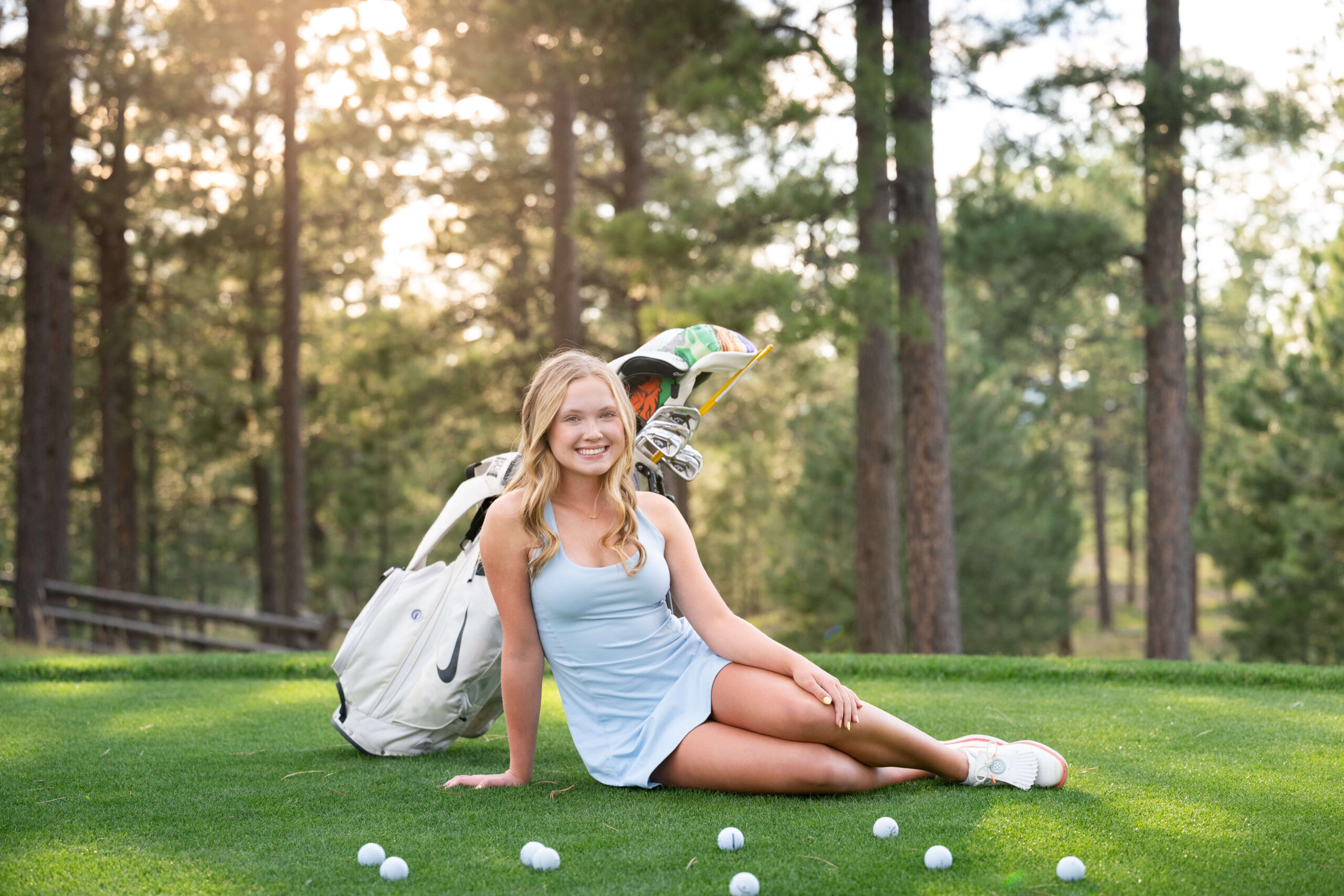 A high school senior girl in a blue dress sits on a putting green at sunset with her golf clubs behind her after finding teenage volunteer opportunities in colorado springs.